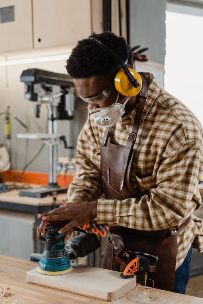 African American craftsman sanding wood in a workshop setting wearing safety gear.
